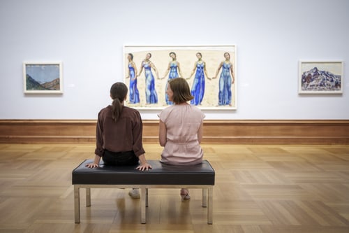 Zwei Personen sitzen auf einer Bank in einer Kunstgalerie, umgeben von Gemälden.