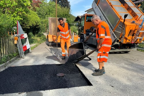 Zwei Personen in orangefarbener Arbeitskleidung reparieren eine Strasse.