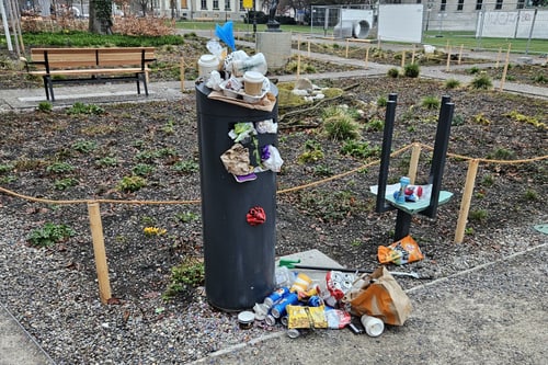 Überfüllter Mülleimer in einem Park mit verstreutem Müll.