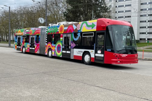 Bunter Gelenkbus auf leerer Strasse vor Gebäuden.