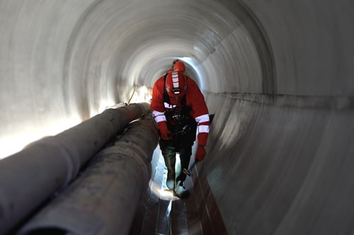 Person in roter Schutzkleidung kriecht durch einen Tunnel mit Rohren.