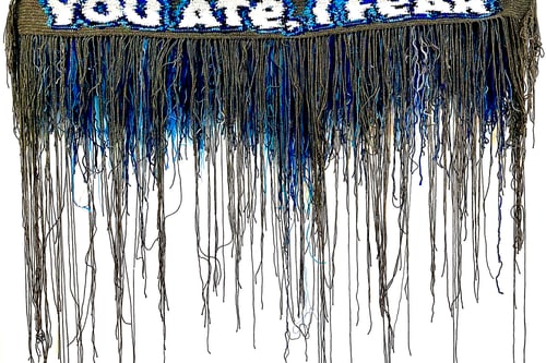 Hängendes Textilkunstwerk mit Fransen und Text.