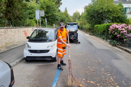 Person in orange Kleidung fegt Blätter auf der Strasse.