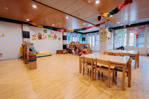 Bunter Kindergartenraum mit Holztischen und Spielbereich.