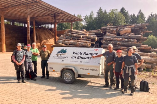 Gruppe von Menschen neben einem Anhänger mit der Aufschrift Wintiranger im Einsatz vor gestapelten Baumstämmen.