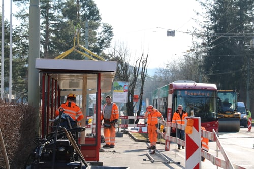 Bauarbeiter in oranger Kleidung bei Bushaltestellenbau, Bus im Hintergrund.