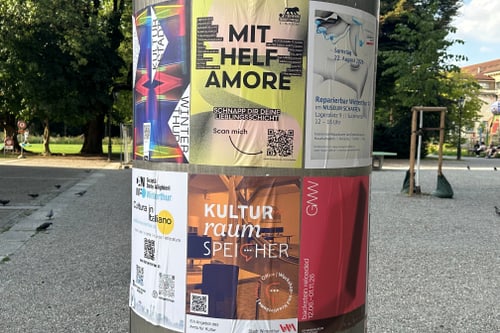 Runde Werbesäule mit bunten Plakaten auf einem Platz.
