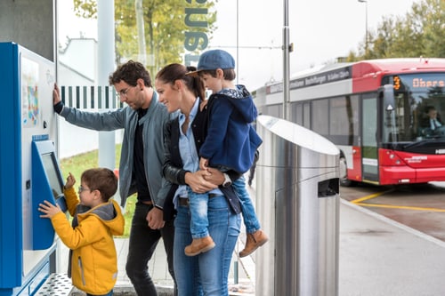 Familie kauft Ticket am Automaten mit Bus im Hintergrund.