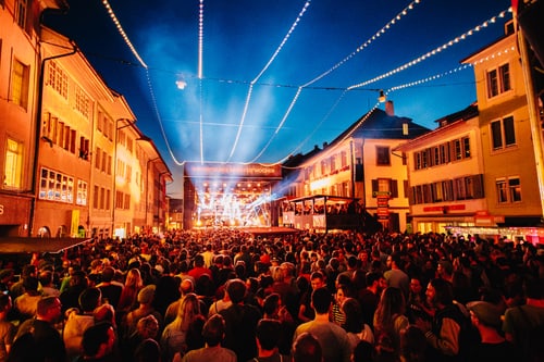 Menschenmenge auf einem Festivalplatz bei Nacht mit bunten Lichtern.