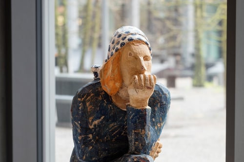 Holzskulptur einer nachdenklichen Person sitzt auf einem Holzhocker vor einem Fenster.