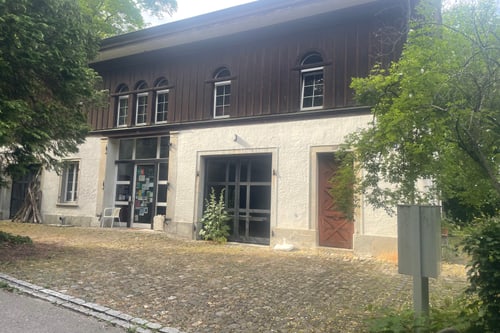 Zweistöckiges Gebäude mit Holzwänden und gepflastertem Vorplatz, umgeben von Bäumen.