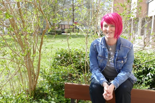 Person mit pinken Haaren sitzt auf einer Bank im Garten.
