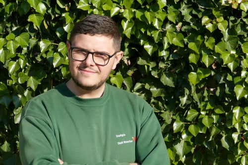 Person mit Brille vor einer grünen Blätterwand.
