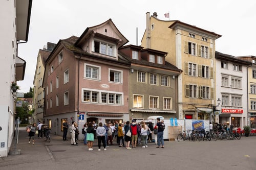 Gruppe von Menschen steht auf einem Platz vor mehrstöckigen Gebäuden.