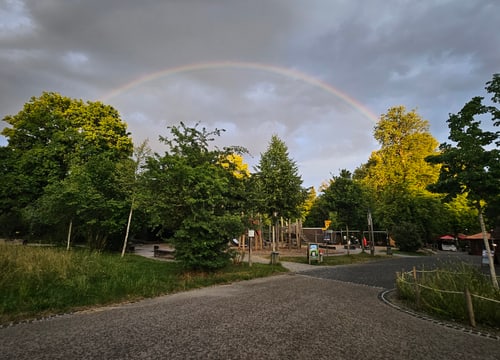 Regenbogen über dem Wildpark Bruderhaus mit Bäumen und Menschen.