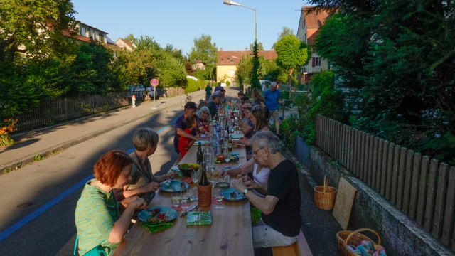 Menschen sitzen an einem langen Tisch auf einer Strasse und essen.