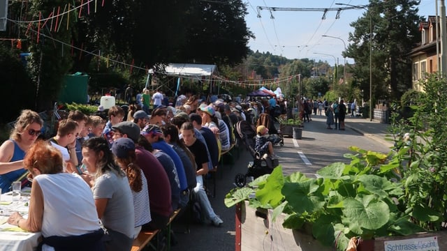 Strassenszene mit vielen Menschen, die an langen Tischen essen.