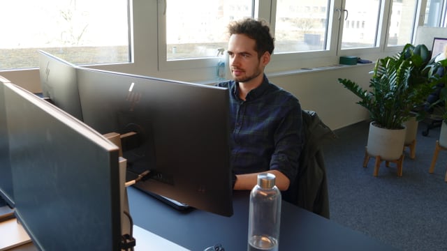 Person am Schreibtisch mit zwei Monitoren und einer Wasserflasche.