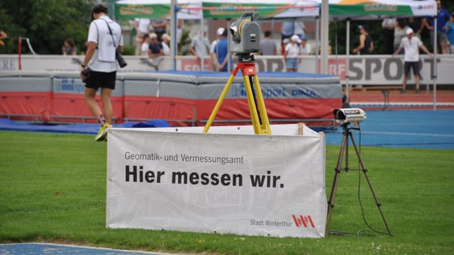 Messgerät auf einem Stativ auf einem Sportplatz mit Menschen im Hintergrund.
