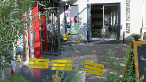 Gelbe Bistrostühle vor einem Gebäudeeingang mit Schildern und Pflanzen.