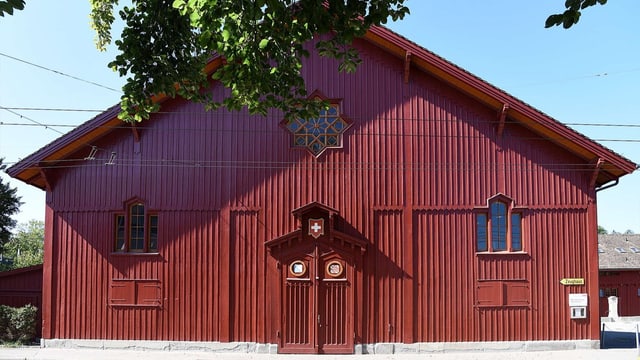 Rotes Holzgebäude mit Fenstern und Baum im Vordergrund.