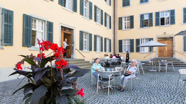 Menschen sitzen auf einem gepflasterten Hof mit Tischen und einem grossen Gebäude im Hintergrund.