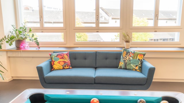 Ein blauer Sofa mit bunten Kissen vor Fenstern, im Vordergrund ein Billardtisch.