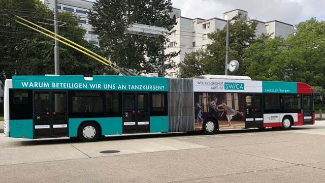 Gelenkbus mit Werbung, Gebäude und Bäume im Hintergrund.