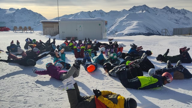Personengruppe liegt im Schnee auf einem Berg mit Winterlandschaft im Hintergrund.