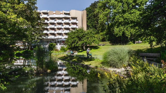 Mehrstöckiges Gebäude vor einem Teich mit Bäumen und einer Person auf dem Rasen.