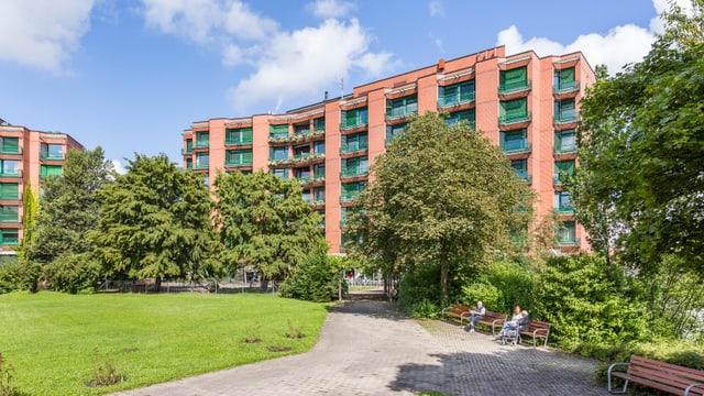 Mehrstöckiges rotes Gebäude hinter einem Park mit Bäumen und Bänken.