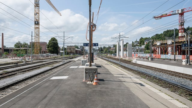 Leerer Bahnsteig mit Gleisen, Kräne im Hintergrund.