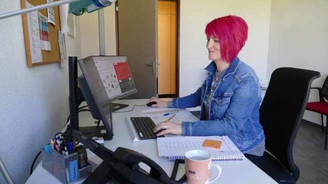 Person mit pinken Haaren arbeitet an einem Computer in einem Büro.