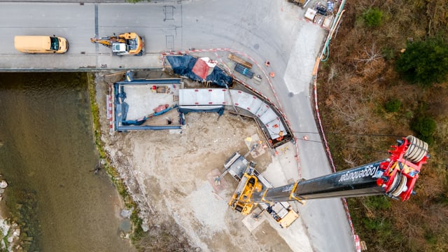 Luftaufnahme einer Baustelle mit einem Kran und einem Lastwagen auf einer Strasse neben einem Fluss.