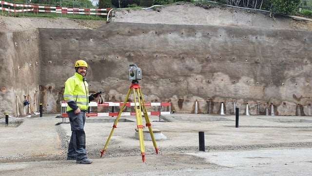 Bauarbeiter mit gelbem Helm neben Vermessungsgerät vor Erdwand.