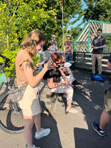 Gruppe von Menschen im Freien, einige sitzen, andere stehen, bei sonnigem Wetter.