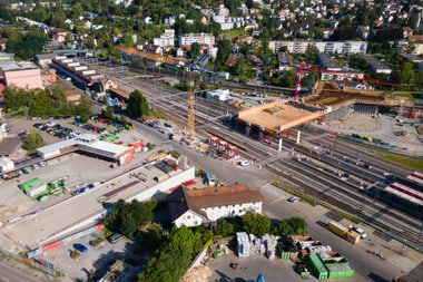 Luftaufnahme einer Baustelle mit Gleisen und Gebäuden in einem städtischen Gebiet.