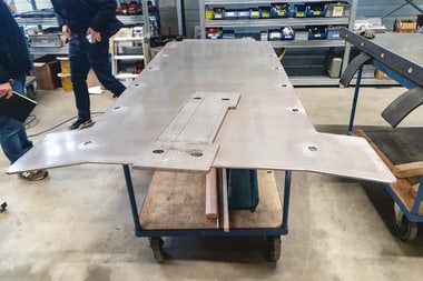 Metallplatte auf Werkbank in Werkstatt mit Personen im Hintergrund.