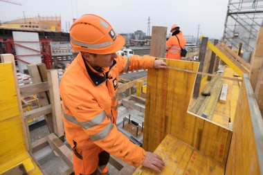 Zwei Bauarbeiter in orangefarbenen Sicherheitskleidung auf einer Baustelle.