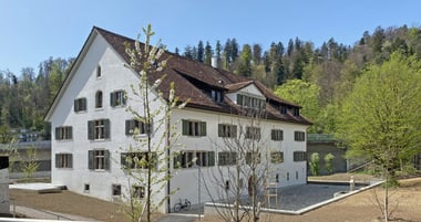 Grosses weisses Gebäude mit Dachschindeln vor einem bewaldeten Hang.