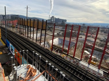 Baustelle neben Bahngleisen mit Industriegelände im Hintergrund.