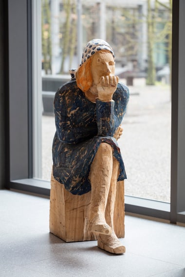 Holzskulptur einer nachdenklichen Person sitzt auf einem Holzhocker vor einem Fenster.