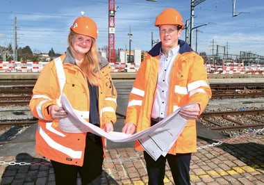 Zwei Personen in orangefarbener Kleidung mit Bauhelmen halten Pläne vor Bahngleisen.