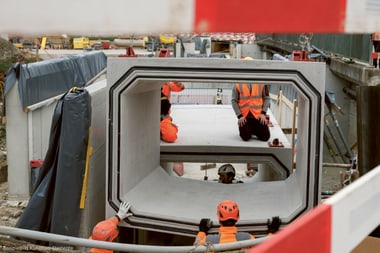 Bauarbeiter mit orangefarbener Schutzkleidung arbeiten an grossen Betonelementen.